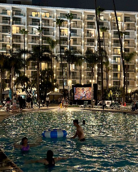 The image shows a night scene at a swimming pool, where people are enjoying the water while a movie is being projected on a screen in the background.