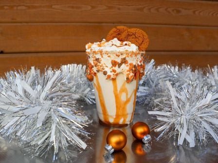 A milkshake topped with whipped cream and cookies, surrounded by silver tinsel and two small orange ornaments, sits on a shiny surface.