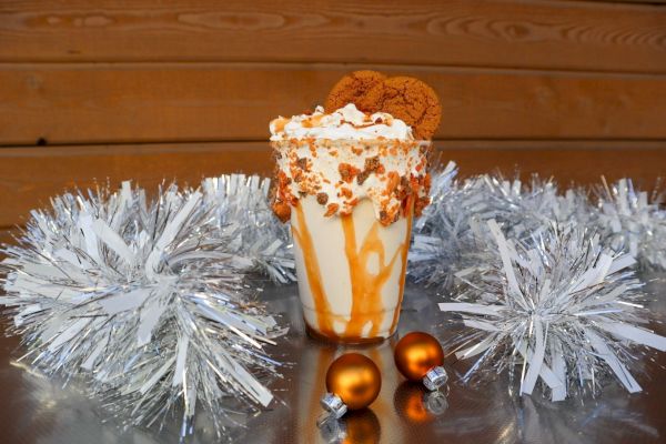 A milkshake topped with whipped cream and cookies, surrounded by silver tinsel and two small orange ornaments, sits on a shiny surface.