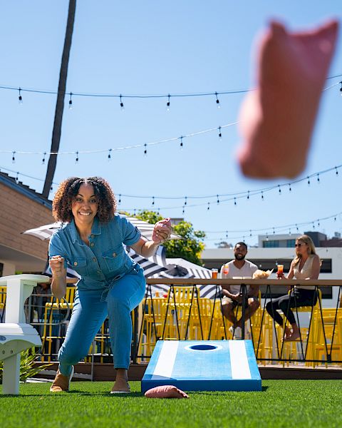 A person playing cornhole outdoors on a sunny day, with others sitting in the background. Hanging string lights add to the festive atmosphere.