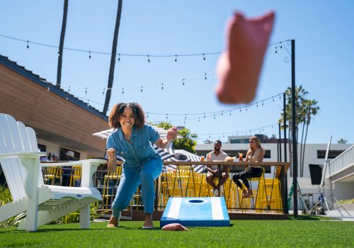 A person playing cornhole outdoors on a sunny day, with others sitting in the background. Hanging string lights add to the festive atmosphere.