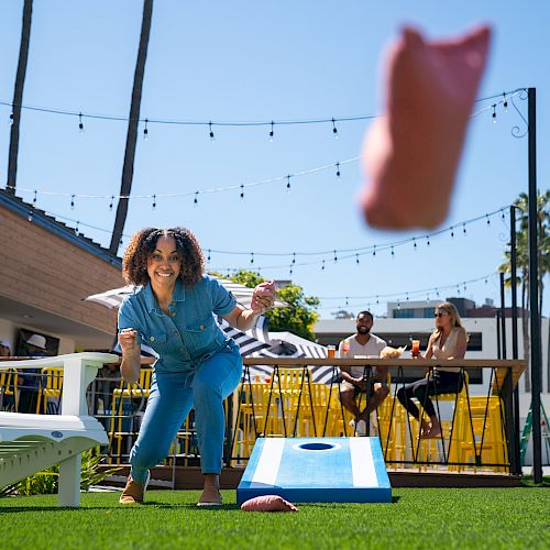 A person playing cornhole outdoors on a sunny day, with others sitting in the background. Hanging string lights add to the festive atmosphere.