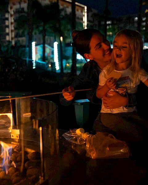 A person and a child are roasting marshmallows over a fire pit at night, with a box of S'mores ingredients nearby.