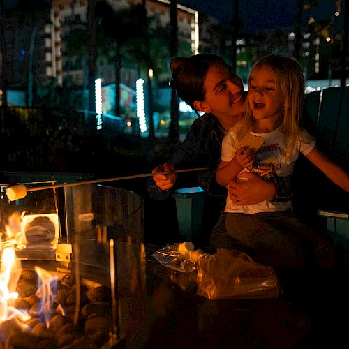 A person and a child are roasting marshmallows over a fire pit at night, with a box of S'mores ingredients nearby.