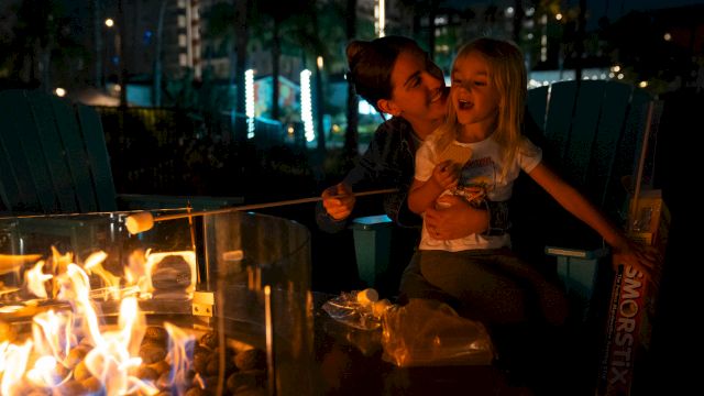 A person and a child are roasting marshmallows over a fire pit at night, with a box of S'mores ingredients nearby.