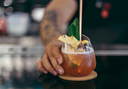 A hand with a tattoo is holding a cocktail garnished with pineapple and a green leaf on a coaster, sitting on a dark countertop.