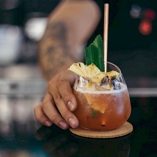 A hand with a tattoo is holding a cocktail garnished with pineapple and a green leaf on a coaster, sitting on a dark countertop.