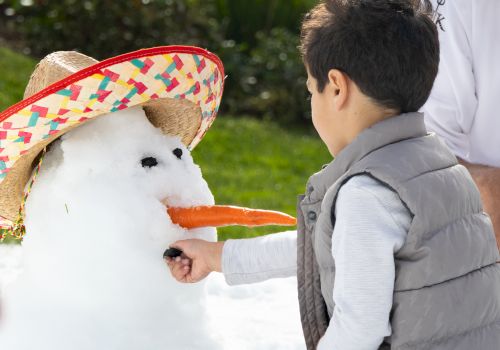 A child places a button on a snowman with a carrot nose and a sombrero hat, while a person in white appears partially in the background.