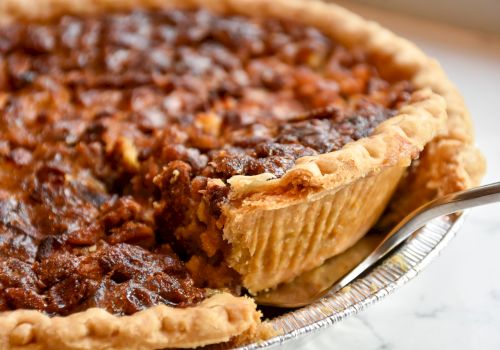 The image shows a pecan pie with a slice being taken out, revealing its nutty filling and flaky crust in a metal pie tin.