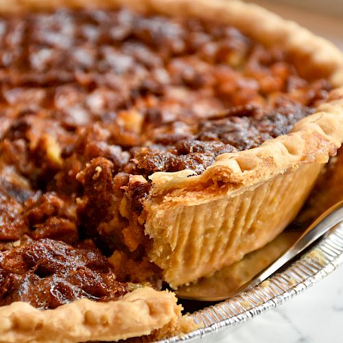 The image shows a pecan pie with a slice being taken out, revealing its nutty filling and flaky crust in a metal pie tin.