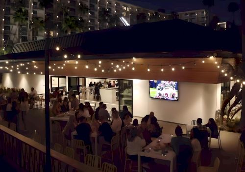 An outdoor dining area at night with string lights, tables of people, and a TV screen showing a sports game.