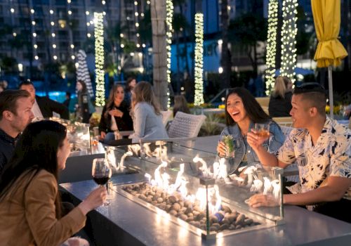 People are enjoying drinks around a fire pit at an outdoor gathering in a lively, illuminated setting.