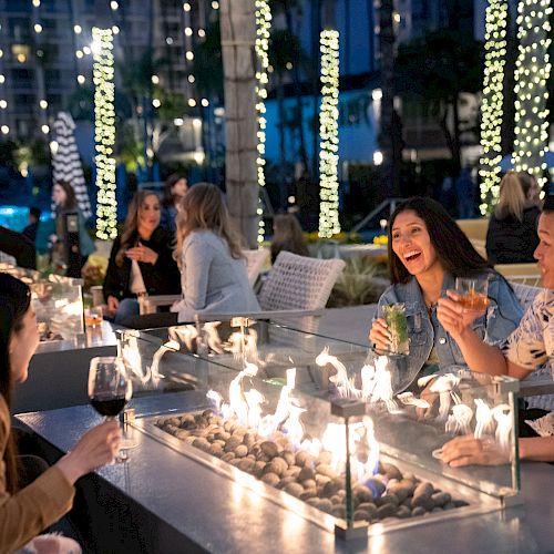 People are enjoying drinks around a fire pit at an outdoor gathering in a lively, illuminated setting.