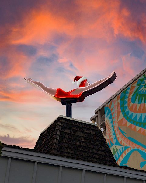 A rooftop sculpture of a swimsuit-clad diver against a colorful sunset sky, with a building showing a geometric mural in the background.