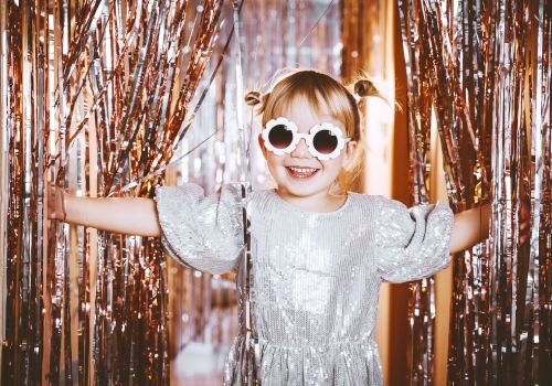 A child in a sparkly dress and sunglasses smiles, surrounded by metallic streamers, creating a festive and joyful scene.