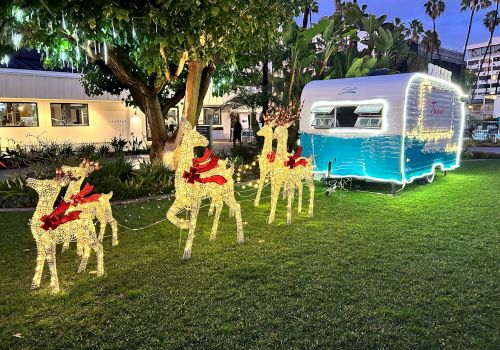 The image shows illuminated reindeer decorations with red bows on grass, next to a blue and white trailer, under a large tree at night.