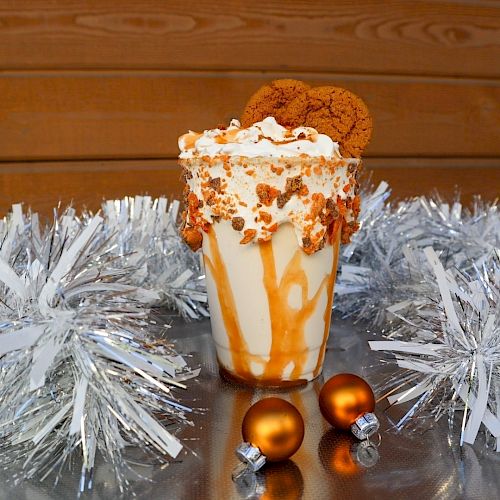 A caramel milkshake topped with whipped cream, cookies, and caramel drizzle, surrounded by silver tinsel and small gold ornaments.