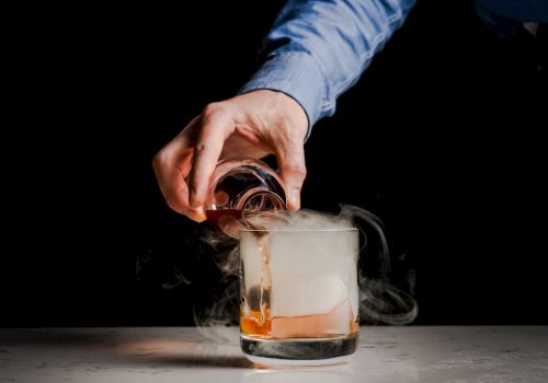 A hand pours a liquid into a glass with a large ice cube, creating smoke or mist, likely from dry ice or a smoky effect.