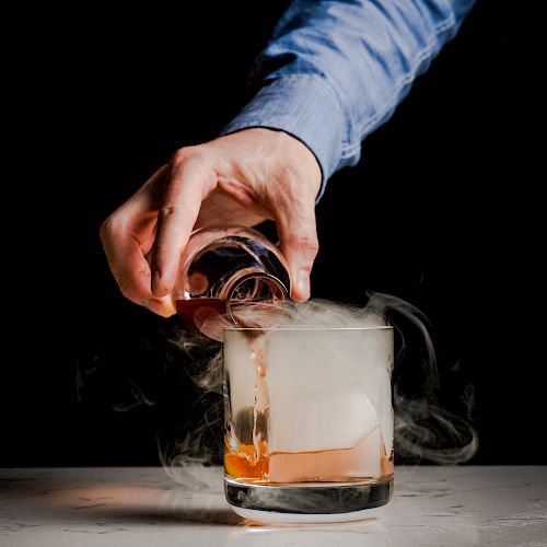 A hand pours a liquid into a glass with a large ice cube, creating smoke or mist, likely from dry ice or a smoky effect.