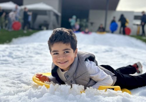 A child is lying on snow with a sled, smiling, in front of a modern building.