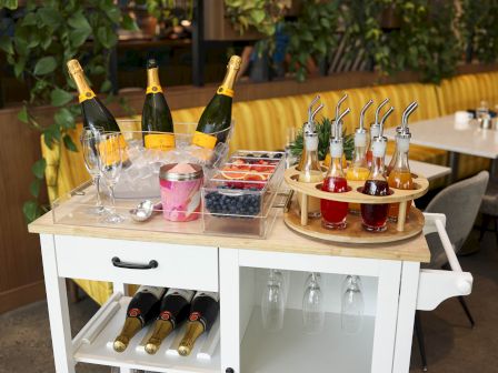 A beverage cart with champagne bottles, glasses, fruit trays, and colorful syrups is set up near a dining area with green plants.