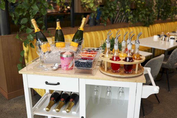 A beverage cart with champagne bottles, glasses, fruit trays, and colorful syrups is set up near a dining area with green plants.