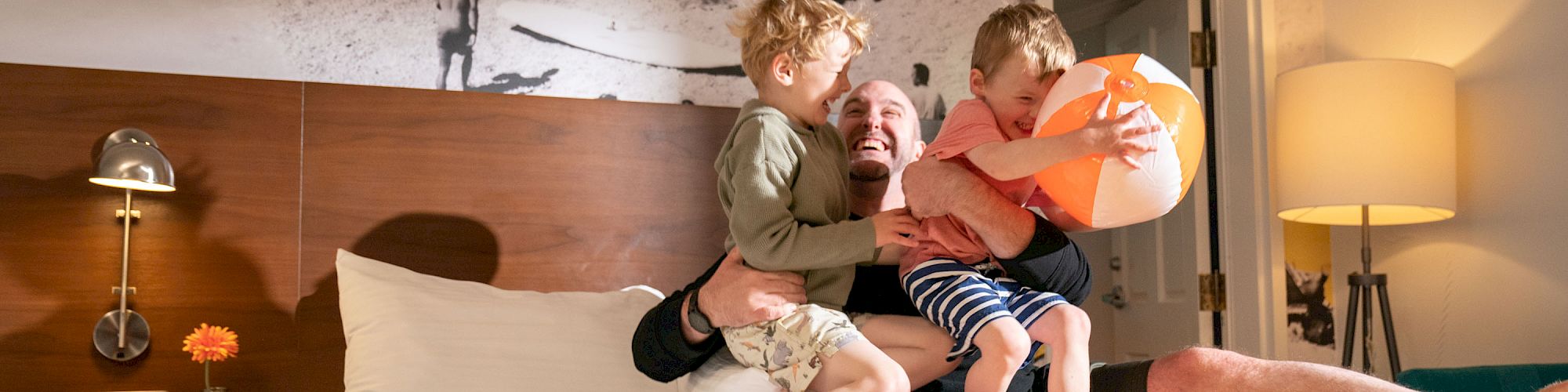 A person and two kids are joyfully playing on a bed in a cozy room, with a beach-themed wall mural and a warm yellow throw.