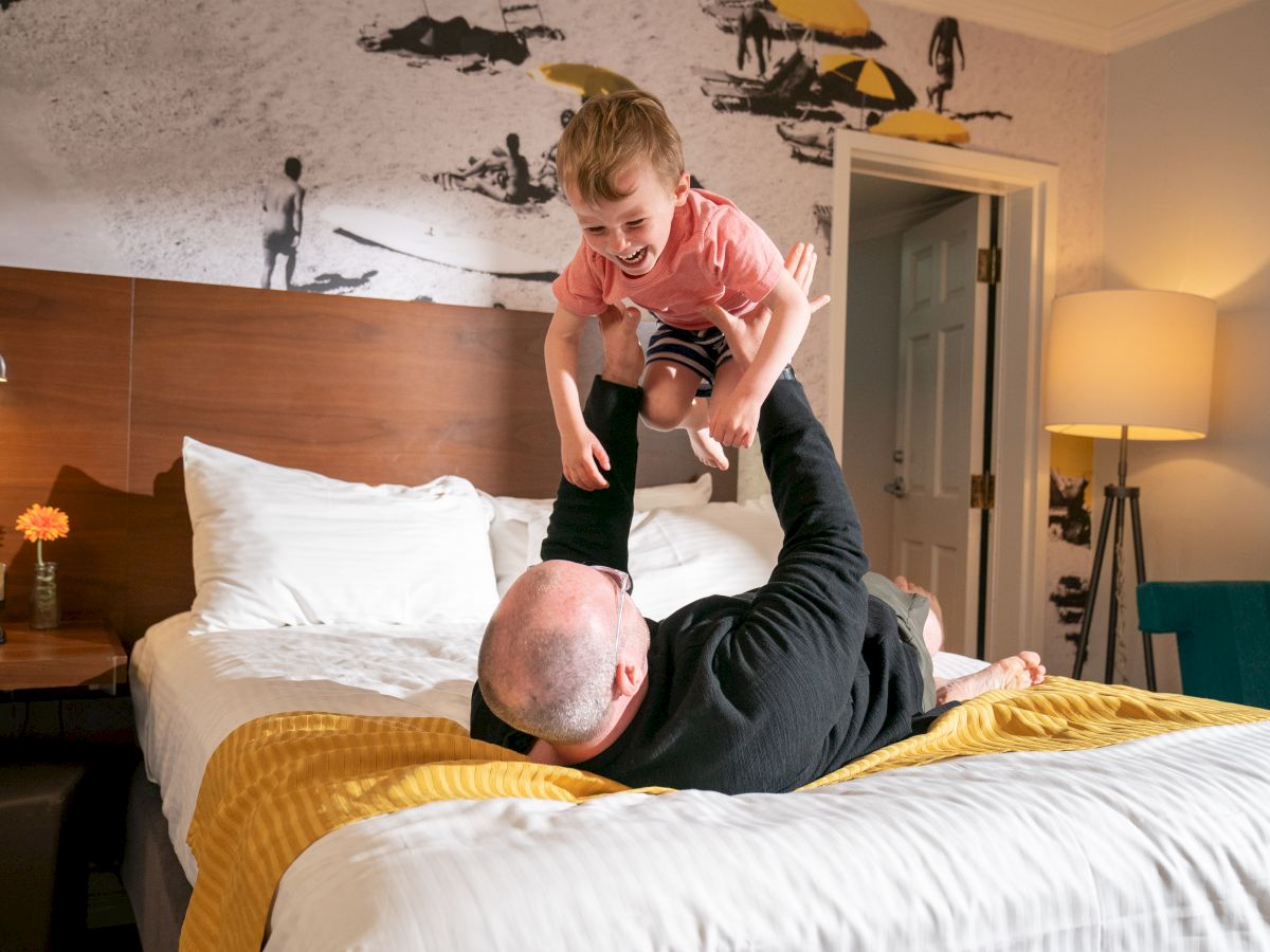 A child is joyfully being lifted in the air by an adult lying on a bed, in a cozy room with a beach-themed wall mural and warm lighting.