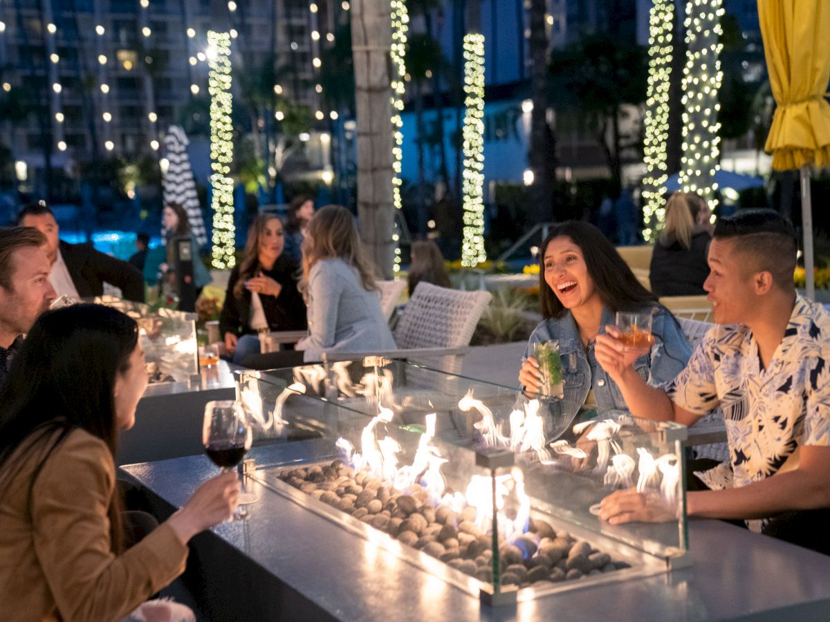 A group of people is enjoying drinks and conversation around a lit fire table outdoors, with string lights and a festive ambiance.