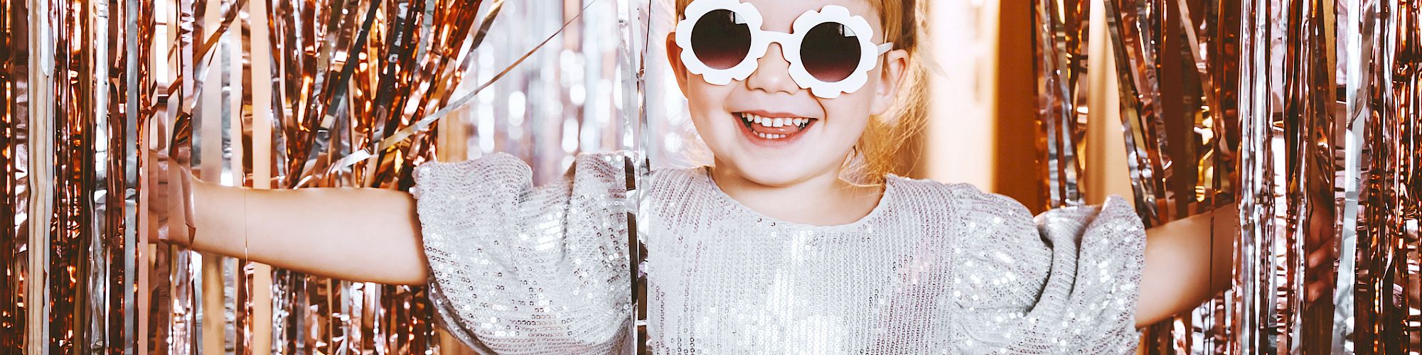 A child in a shiny silver dress and round sunglasses stands between sparkly fringe curtains, smiling widely.