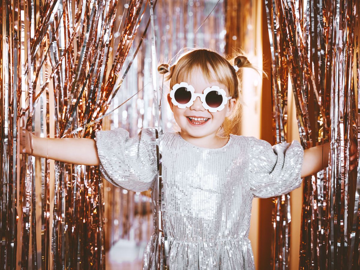 A child in a shiny dress and round sunglasses stands joyfully between metallic streamers, creating a festive and playful atmosphere.