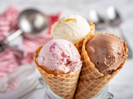 Three waffle cones filled with scoops of vanilla, strawberry, and chocolate ice cream sit together, with a blurred background of utensils.