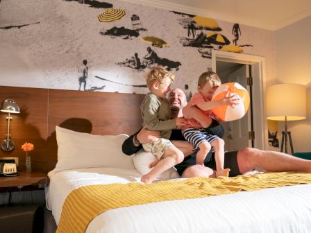 A joyful moment in a hotel room features three people playing on a bed with a beach ball, against a backdrop of a beach-themed decor wall.