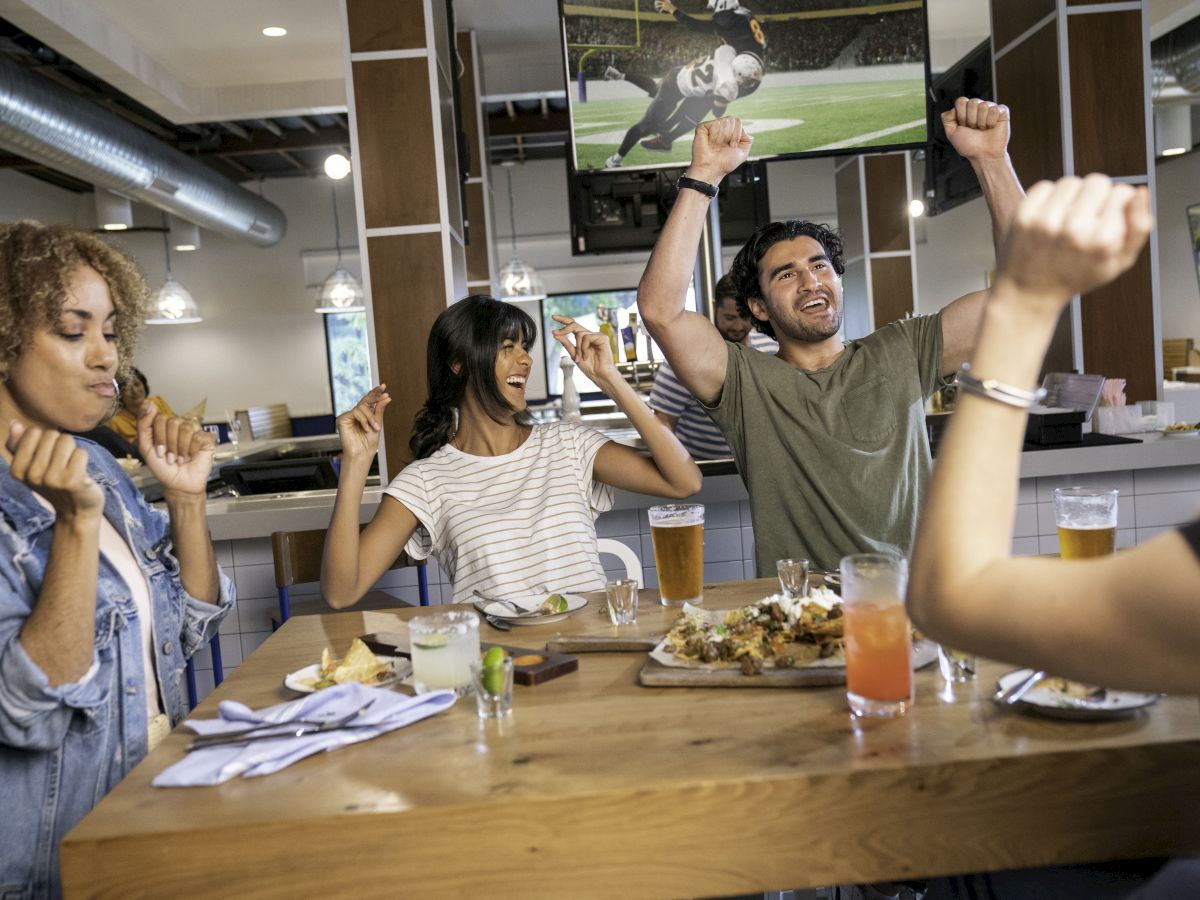 A group of people cheer and enjoy food and drinks while watching a sports game on TV in a lively setting.