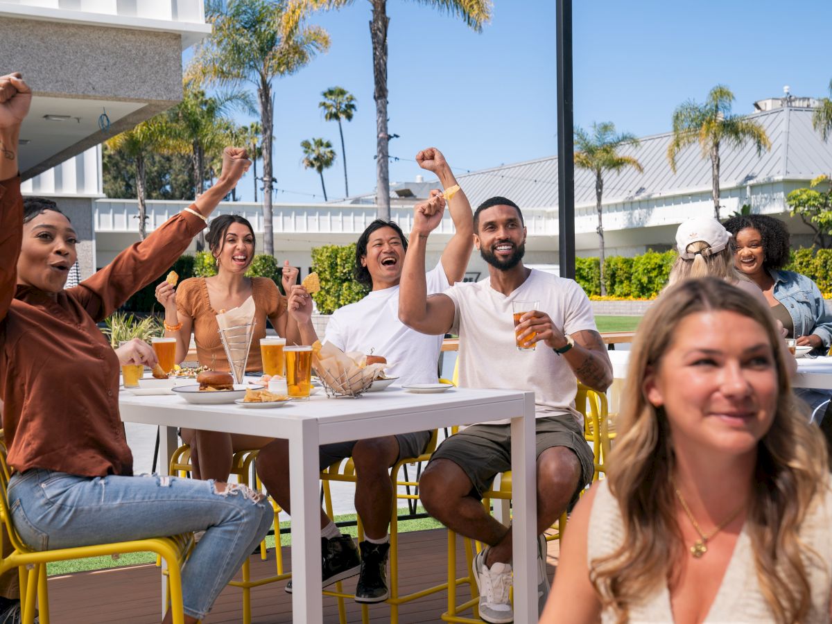 A group of people are smiling and cheering at an outdoor gathering with drinks and food on the table.