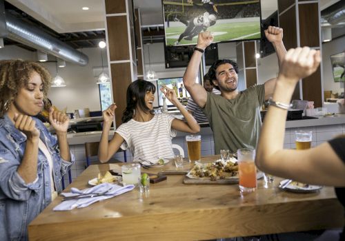 People are cheering and eating at a table in a restaurant with a sports game on TV, enjoying drinks and food.