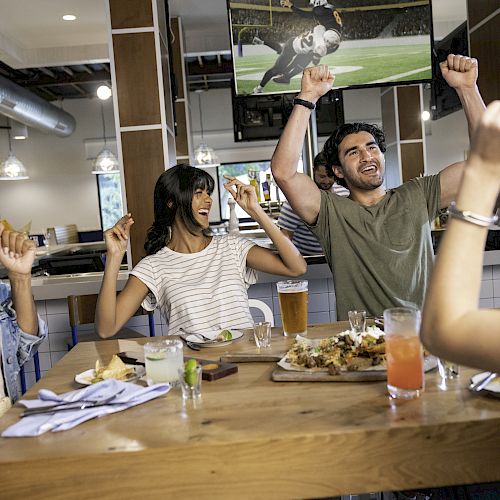 People are cheering and eating at a table in a restaurant with a sports game on TV, enjoying drinks and food.