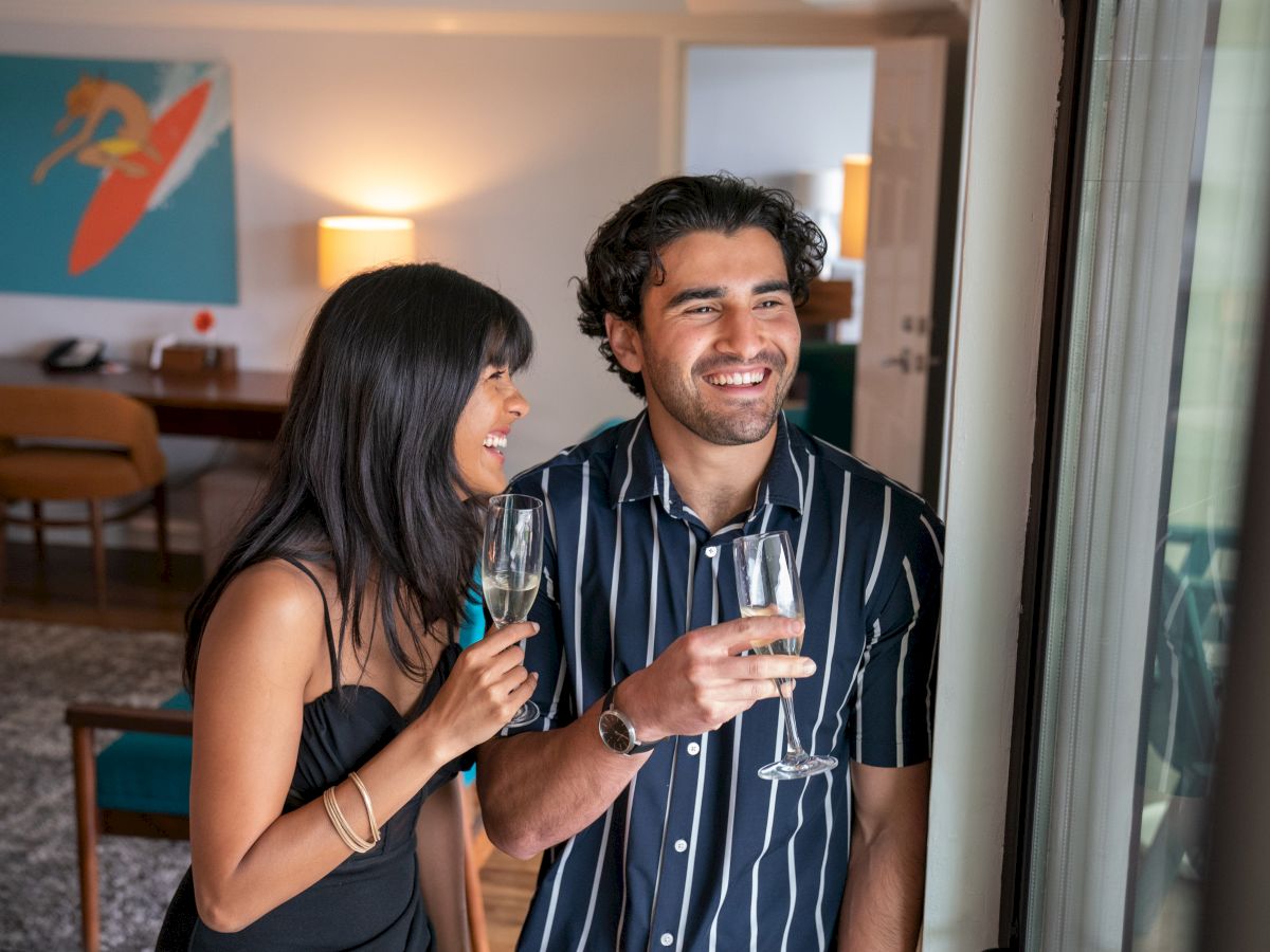 A couple is indoors, smiling and holding champagne glasses, standing near a window. There’s a colorful painting in the background.