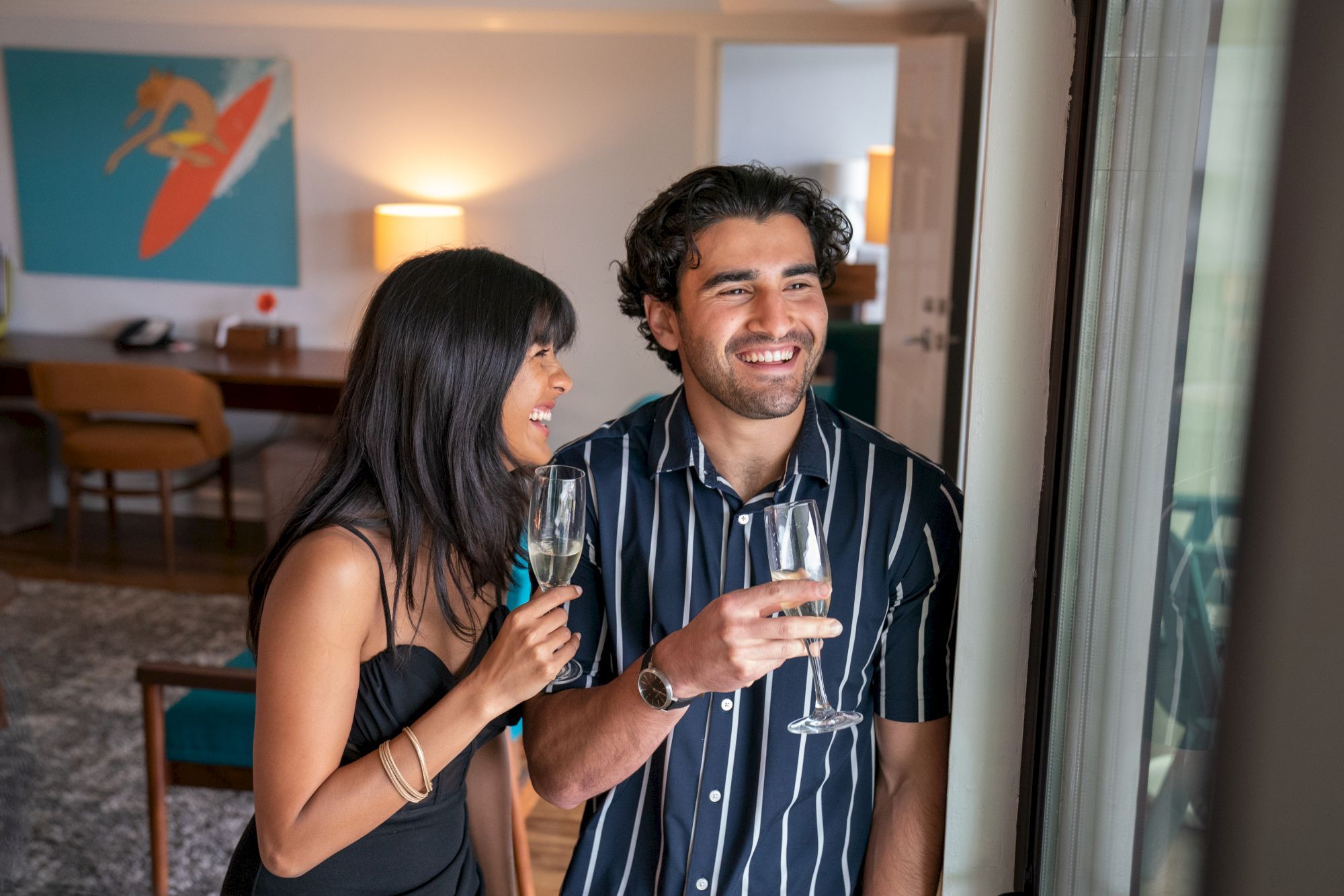 A couple is standing indoors, smiling, holding champagne glasses, with artwork and furniture in the background.