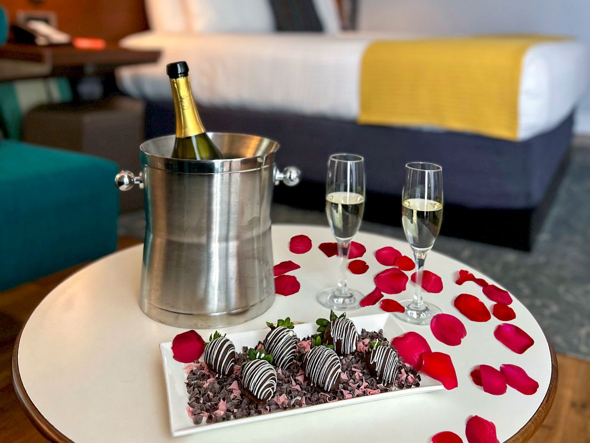 A hotel room with a bed, champagne in an ice bucket, two glasses, chocolate-covered strawberries, and rose petals on a table.