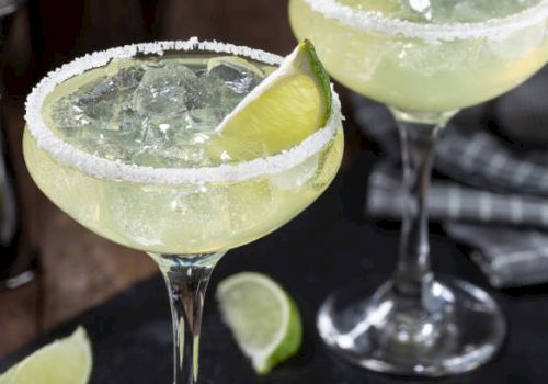 Two margarita cocktails with salted rims, ice, and lime wedges, served in elegant glasses. Limes are also scattered on the table nearby.