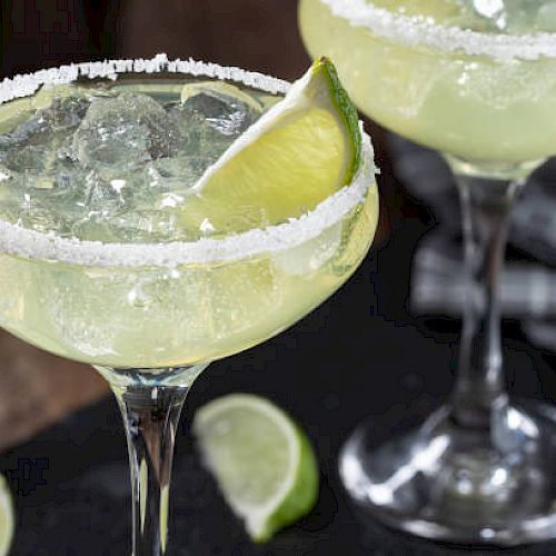 Two margarita cocktails with salted rims, ice, and lime wedges, served in elegant glasses. Limes are also scattered on the table nearby.