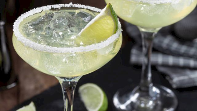 Two margarita cocktails with salted rims, ice, and lime wedges, served in elegant glasses. Limes are also scattered on the table nearby.