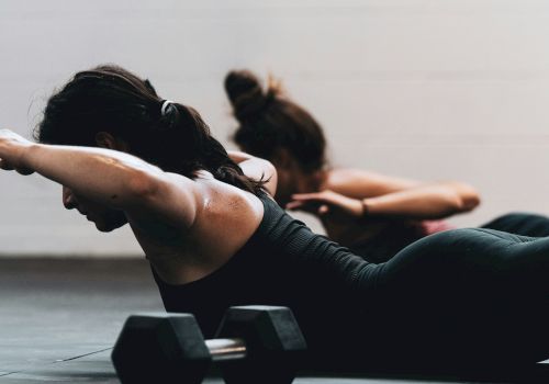 Two individuals on a gym floor perform exercises with arms extended, facing down, near a pair of dumbbells.