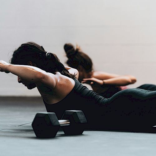 Two individuals on a gym floor perform exercises with arms extended, facing down, near a pair of dumbbells.