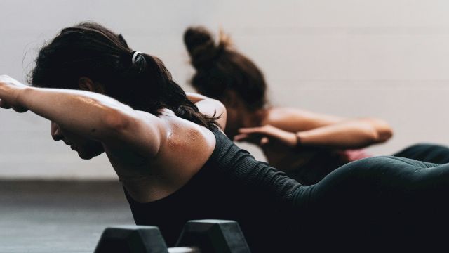 Two individuals on a gym floor perform exercises with arms extended, facing down, near a pair of dumbbells.