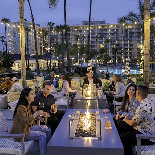 People are gathered around outdoor fire pits, enjoying drinks under string lights, with palm trees and a hotel in the background.