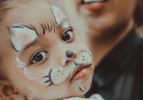 A child with a cat face paint looks at the camera, with an adult in the background.