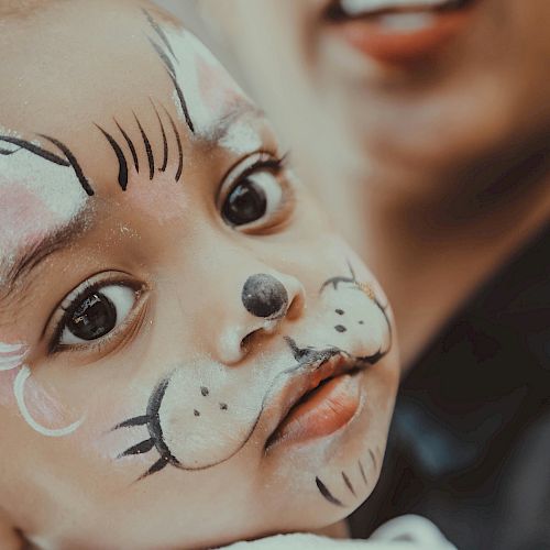 A child with a cat face paint looks at the camera, with an adult in the background.