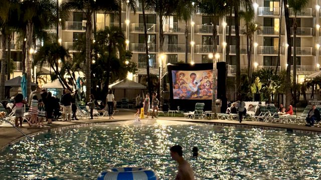 The image shows a nighttime scene with people swimming in a pool while a movie plays on a screen beside the pool, surrounded by lit buildings.